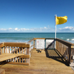 Yellow beach flag blows in the wind at the end of a wooden dune crossover where a sign depicts the meaning of the flag colors