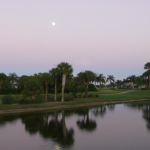 A view across a calm and reflective lake to the golf course as the full moon shines in a milky evening sky. 