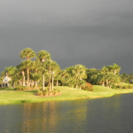 A stormy sky is ominously reflected in a calm lake, as the landscaping and homes next to the lake are bathed in gold sunlight. 