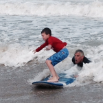 A boy stands carefully balanced atop a boogie board as he rides a wave onto shore aided by male relative.