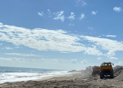 Heavy equipment, engaged in beach repair, drives on the beach alongside the ocean on a sunny day.