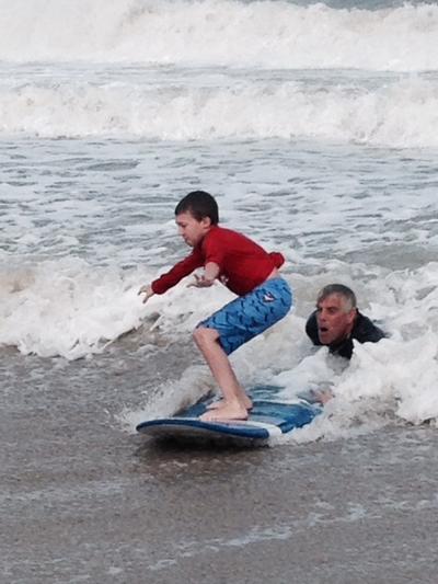 A boy stands carefully balanced atop a boogie board as he rides a wave onto shore aided by male relative.
