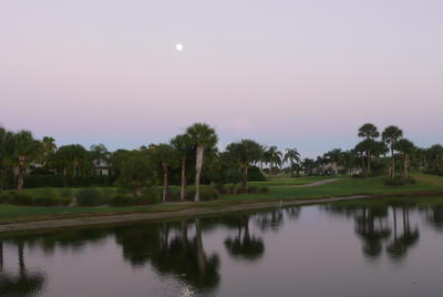 A view across a calm and reflective lake to the golf course as the full moon shines in a milky evening sky. 