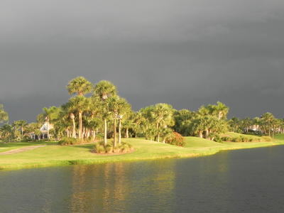 A stormy sky is ominously reflected in a calm lake, as the landscaping and homes next to the lake are bathed in gold sunlight. 